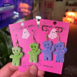May include: Two pairs of earrings featuring gummy bear charms. One pair is green and the other is blue. The earrings are displayed on pink cards with the text "Creepin' it cute" and a ghost illustration.