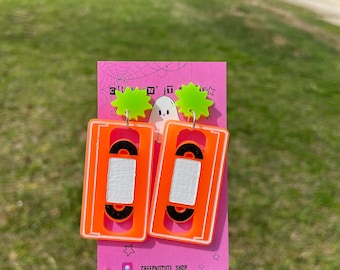 May include: A pair of orange and white cassette tape earrings with neon green starburst accents. The earrings are displayed on a pink card with a ghost graphic and the text "CREEPNITCUTE_SHOP".