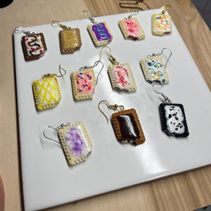 Puede incluir: Una colección de pendientes Pop-Tart hechos a mano con varios diseños de glaseado y relleno. Cada pendiente presenta una forma rectangular de pastel de color marrón dorado con glaseado y chispas coloridas, sujeto a un gancho para la oreja plateado o dorado. Los pendientes se exhiben sobre una superficie blanca.