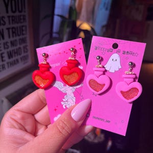 May include: Heart-shaped earrings with a red potion bottle design. The earrings are displayed on pink cards with the text "GREEPIN IT CUTE" and "CREEPNITCUTE_SHOP". They feature gold-coloured accents and a glittery centre.