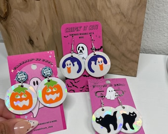 Könnte beinhalten: Halloween-Ohrringe mit verschiedenen Designs, darunter Kürbisse, Geister und schwarze Katzen. Die Ohrringe sind rund und haben holografische Akzente. Jedes Paar wird auf einer rosa Karte mit dem Text "Creepin' It Cute" präsentiert.