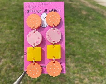 May include: A pair of earrings with a pink, yellow, and orange color scheme. The earrings feature a cookie-shaped charm, a square charm, and a round charm. The earrings are on a pink background with the text "Creepin' it Cute" and a ghost.
