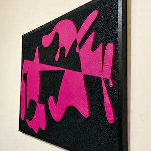 May include: Abstract wall art featuring a black textured background with bright pink, raised shapes. The artwork is framed in a black wooden frame, creating a modern and bold aesthetic. The shapes appear to be dripping or flowing.