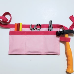 Pale pink cotton toolbelt for small children, with three large pockets and dark pink trim and ties. There is an added loop off to one side for hanging a small tool, such as a hammer. This view shows the pockets and hanging loop filled with toy tools.
