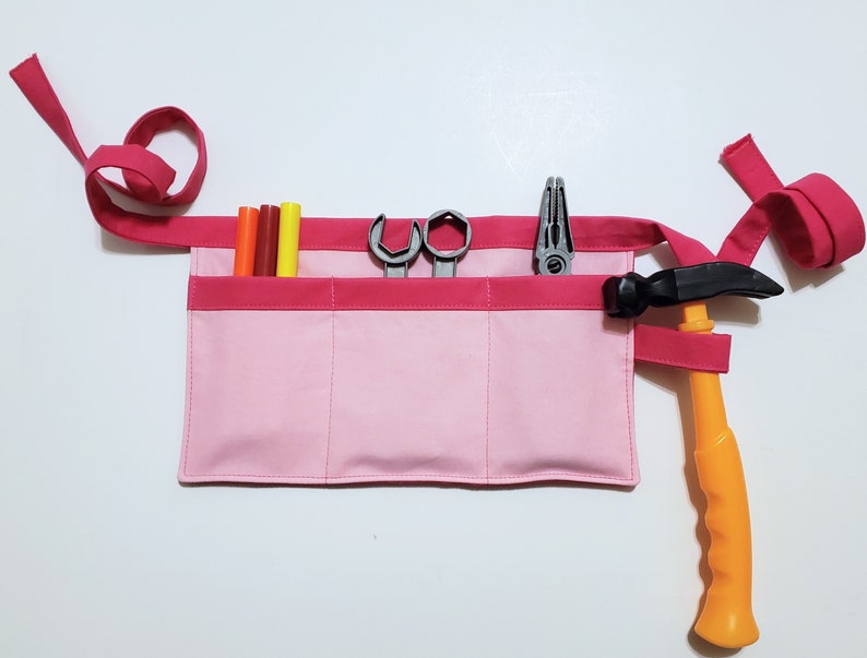Pale pink cotton toolbelt for small children, with three large pockets and dark pink trim and ties. There is an added loop off to one side for hanging a small tool, such as a hammer. This view shows the pockets and hanging loop filled with toy tools.