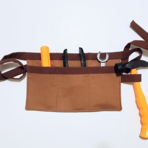 Brown cotton toolbelt for small children, with three large pockets and dark brown trim and ties. There is an added loop off to one side for hanging a small tool, such as a hammer. This view shows the pockets and hanging loop filled with toy tools.
