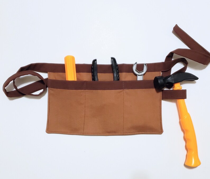 Brown cotton toolbelt for small children, with three large pockets and dark brown trim and ties. There is an added loop off to one side for hanging a small tool, such as a hammer. This view shows the pockets and hanging loop filled with toy tools.