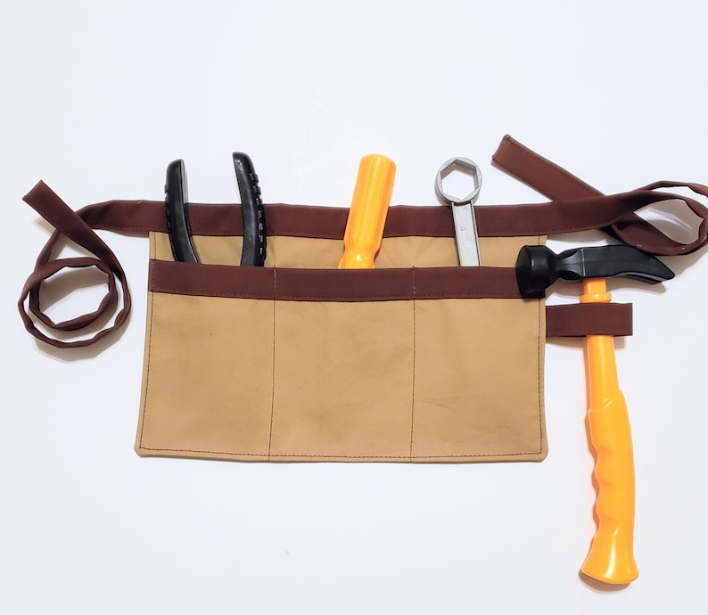 Tan cotton toolbelt for small children, with three large pockets and brown trim and ties. There is an added loop off to one side for hanging a small tool, such as a hammer. This view shows the pockets and hanging loop filled with toy tools.