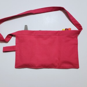 Pale pink cotton toolbelt for small children, with three large pockets and dark pink trim and ties. There is an added loop off to one side for hanging a small tool. This view shows the back, which is dark pink to match the trim. Pockets are in use.
