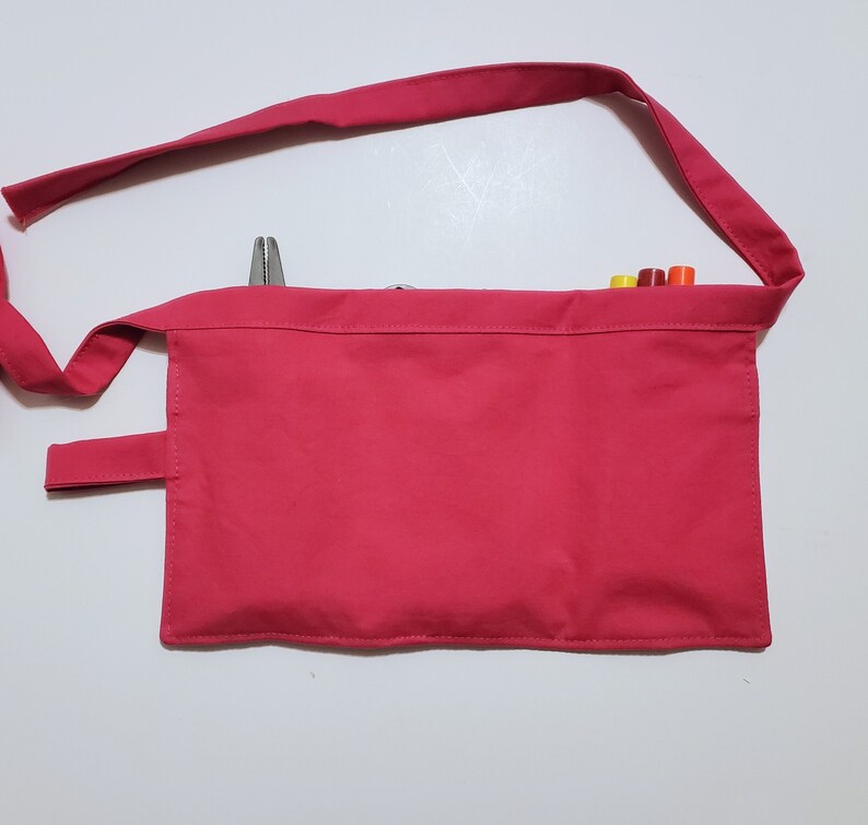 Pale pink cotton toolbelt for small children, with three large pockets and dark pink trim and ties. There is an added loop off to one side for hanging a small tool. This view shows the back, which is dark pink to match the trim. Pockets are in use.