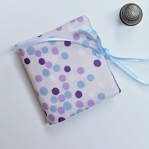May include: A small, rectangular fabric pouch with a light purple background and a pattern of purple, blue, and white polka dots. A light blue ribbon is tied around the top. A silver thimble is next to the pouch.