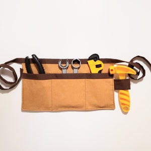 Gold cotton toolbelt for small children, with three large pockets and brown trim and ties. There is an added loop off to one side for hanging a small tool, such as a hammer. This view shows the pockets and hanging loop filled with toy tools.