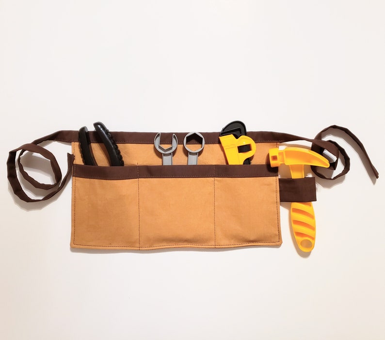 Gold cotton toolbelt for small children, with three large pockets and brown trim and ties. There is an added loop off to one side for hanging a small tool, such as a hammer. This view shows the pockets and hanging loop filled with toy tools.