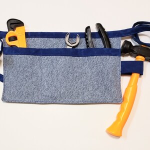 Blue cotton toolbelt for small children, with three large pockets and navy trim and ties. There is an added loop off to one side for hanging a small tool, such as a hammer. This view shows the pockets and hanging loop filled with toy tools.