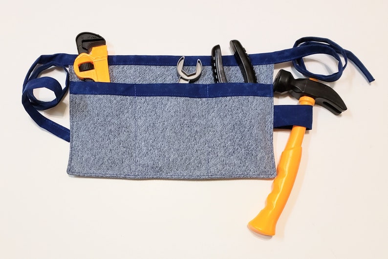 Blue cotton toolbelt for small children, with three large pockets and navy trim and ties. There is an added loop off to one side for hanging a small tool, such as a hammer. This view shows the pockets and hanging loop filled with toy tools.