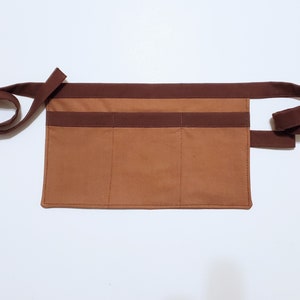 Brown cotton toolbelt for small children, with three large pockets and dark brown trim and ties. There is an added loop off to one side for hanging a small tool, such as a hammer.
