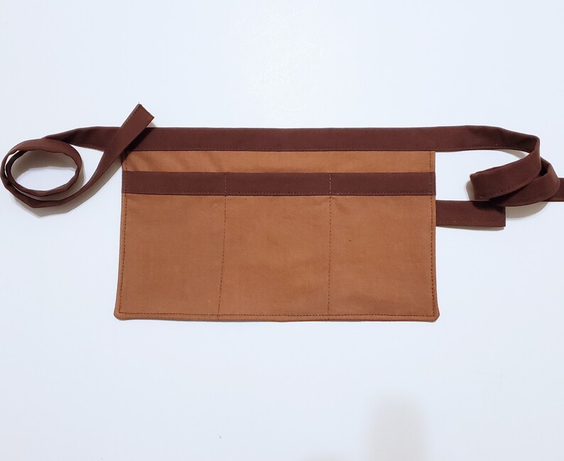 Brown cotton toolbelt for small children, with three large pockets and dark brown trim and ties. There is an added loop off to one side for hanging a small tool, such as a hammer.