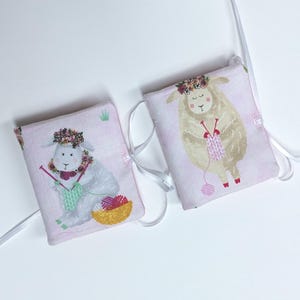 May include: Two rectangular fabric pouches in light pink, each featuring a white ribbon tie. One pouch displays a gray rabbit wearing a floral crown and knitting with green yarn. The other pouch shows a beige sheep with a floral crown knitting.