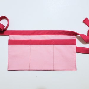 Pale pink cotton toolbelt for small children, with three large pockets and dark pink trim and ties. There is an added loop off to one side for hanging a small tool, such as a hammer.