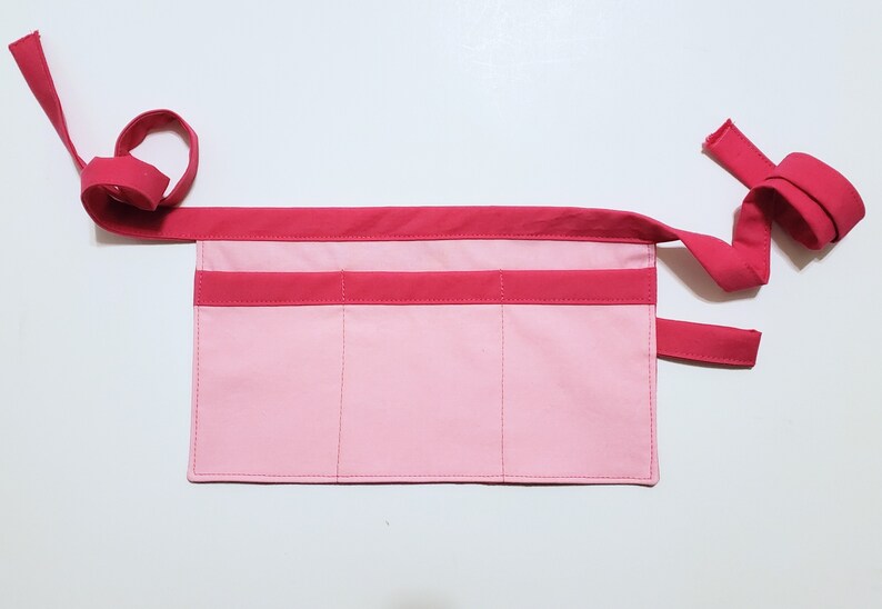 Pale pink cotton toolbelt for small children, with three large pockets and dark pink trim and ties. There is an added loop off to one side for hanging a small tool, such as a hammer.