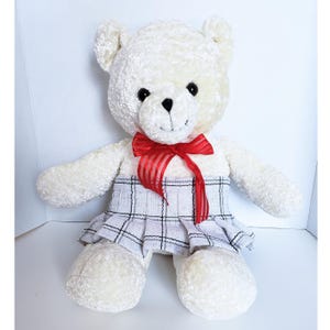 May include: A plush teddy bear with cream-colored fur, featuring black plastic eyes and a stitched nose. The bear wears a bright red sheer ribbon tied in a bow around its neck and a white pleated skirt with a black plaid pattern around its waist.