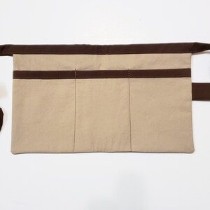 Beige cotton toolbelt for small children, with three large pockets and brown trim and ties. There is an added loop off to one side for hanging a small tool, such as a hammer.