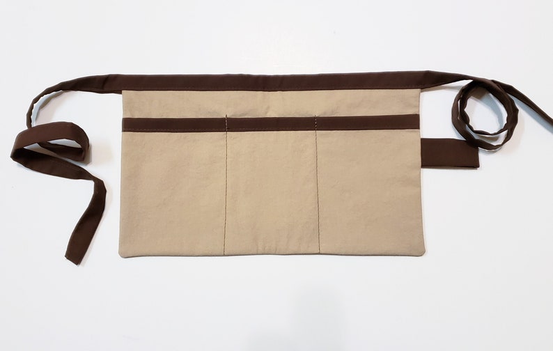 Beige cotton toolbelt for small children, with three large pockets and brown trim and ties. There is an added loop off to one side for hanging a small tool, such as a hammer.