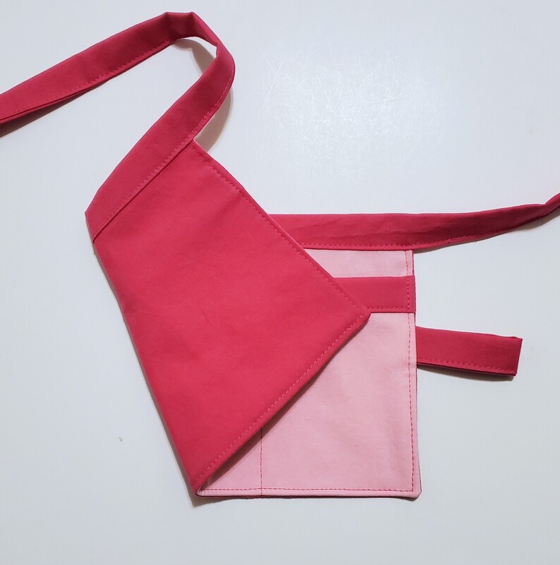 Pale pink cotton toolbelt for small children, with three large pockets and dark pink trim and ties. There is an added loop off to one side for hanging a small tool. This view shows the back, which is dark pink to match the trim.