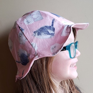 May include: A pink sun hat with a cat print and a wide brim. The hat has a flap that can be pulled down to protect the neck from the sun. The hat is being worn by a young person with long brown hair and blue glasses.