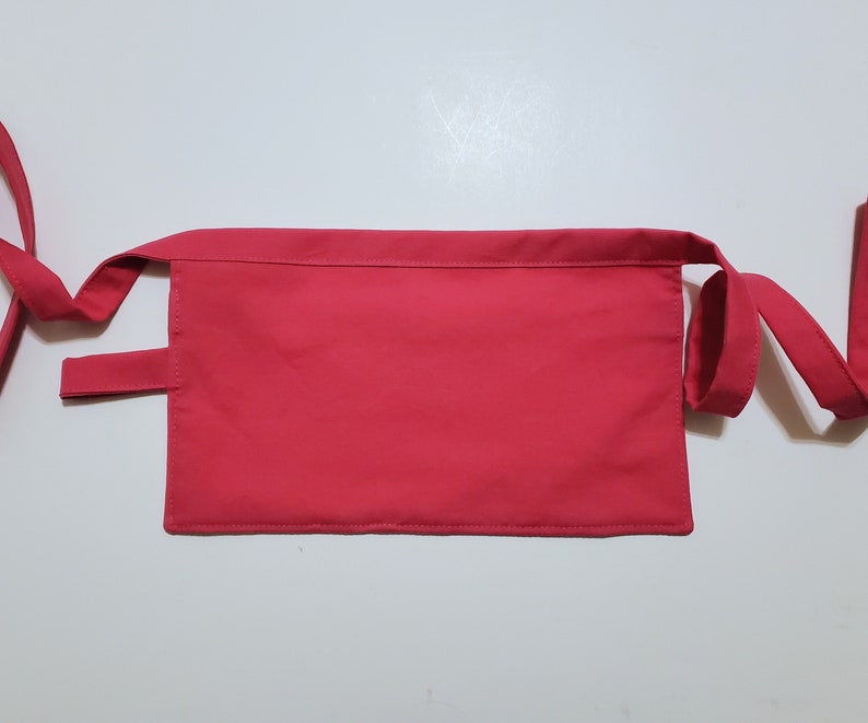 Pale pink cotton toolbelt for small children, with three large pockets and dark pink trim and ties. There is an added loop off to one side for hanging a small tool. This view shows the back, which is dark pink to match the trim.