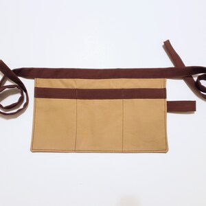 Tan cotton toolbelt for small children, with three large pockets and brown trim and ties. There is an added loop off to one side for hanging a small tool, such as a hammer.