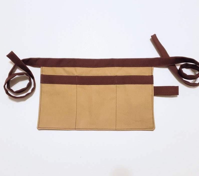 Tan cotton toolbelt for small children, with three large pockets and brown trim and ties. There is an added loop off to one side for hanging a small tool, such as a hammer.