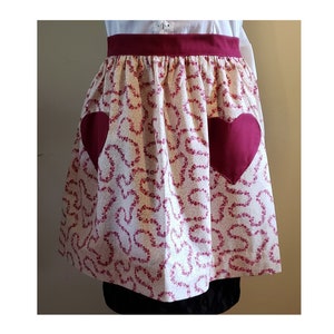 May include: A white apron with a red heart pattern and two large red heart pockets. The apron has a red waistband.