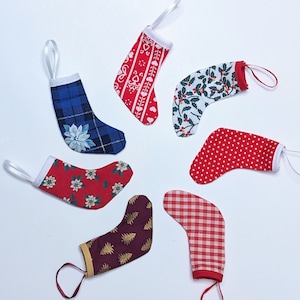 May include: Seven miniature Christmas stockings with white ribbon loops. The stockings display diverse patterns, such as plaid, polka dots, and floral designs, in red, blue, and white. Each stocking is approximately 7.6 cm in length, perfect for festive decorations.