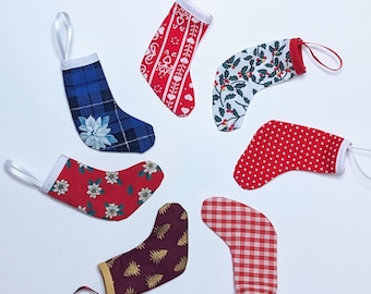 May include: A collection of seven miniature Christmas stockings with white ribbon loops for hanging. The stockings feature various patterns, including plaid, polka dots, floral, and holiday-themed designs in red, blue, and white. Each stocking is approximately 3 inches in length.