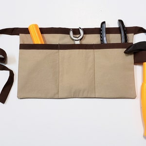 Beige cotton toolbelt for small children, with three large pockets and brown trim and ties. There is an added loop off to one side for hanging a small tool, such as a hammer. This view shows the pockets and hanging loop filled with toy tools.