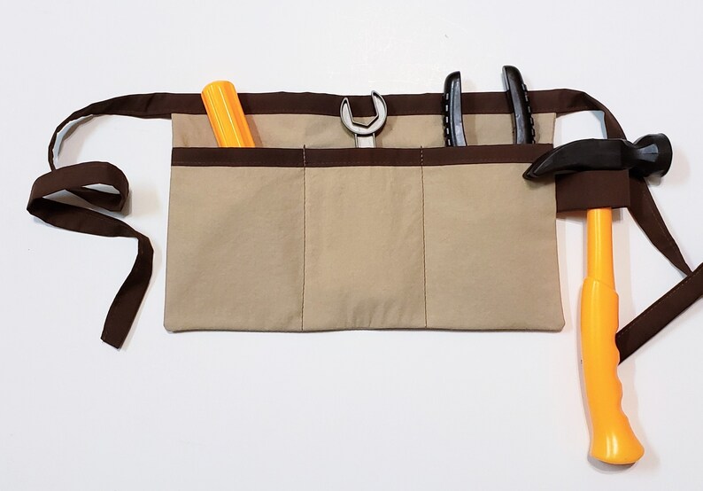 Beige cotton toolbelt for small children, with three large pockets and brown trim and ties. There is an added loop off to one side for hanging a small tool, such as a hammer. This view shows the pockets and hanging loop filled with toy tools.
