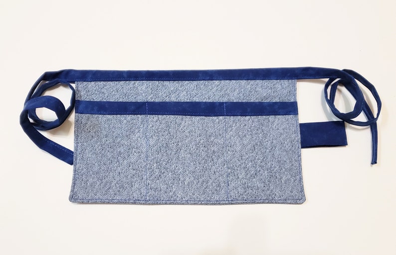 Blue cotton toolbelt for small children, with three large pockets and navy trim and ties. There is an added loop off to one side for hanging a small tool, such as a hammer.