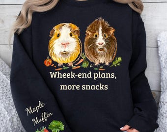 Puede incluir: Una sudadera negra de manga larga con un gráfico de dos cobayas rodeadas de verduras. El texto "Wheek-end plans, more snacks" aparece en el centro, junto con "Maple Muffin" e ilustraciones de una fresa y brócoli en la manga.