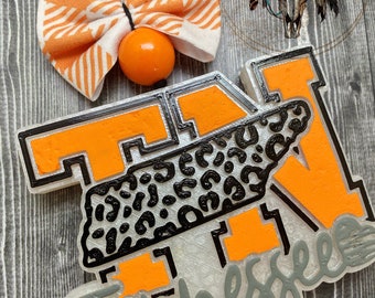May include: A Tennessee car freshie featuring a large orange letter T with a black leopard print overlay, accented by a white and orange plaid fabric bow and a solid orange bead, with the word Tennessee written in grey script at the bottom.