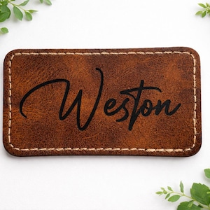 Puede incluir: Un parche rectangular de cuero marrón con el nombre "Weston" en escritura negra. El parche tiene un borde cosido y está rodeado de follaje verde.