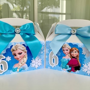K&ouml;nnte beinhalten: Zwei wei&szlig;e Geschenkboxen mit blauen Schleifen und Strassverzierungen. Jede Box zeigt ein Cartoon-Bild von Elsa aus Frozen, Schneeflocken und die Zahl 6. Die Boxen sind mit einem hellblauen und wei&szlig;en Schneeflockenmuster verziert.