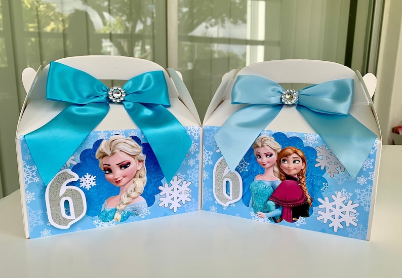 K&ouml;nnte beinhalten: Zwei wei&szlig;e Geschenkboxen mit blauen Schleifen und Strassverzierungen. Jede Box zeigt ein Cartoon-Bild von Elsa aus Frozen, Schneeflocken und die Zahl 6. Die Boxen sind mit einem hellblauen und wei&szlig;en Schneeflockenmuster verziert.