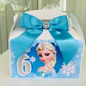 K&ouml;nnte beinhalten: Wei&szlig;e Geschenkbox mit hellblauer Schleife und Strassverzierung. Die Box zeigt Elsa aus Frozen, Schneeflocken und die Zahl 6. Ideal f&uuml;r eine Geburtstagsfeier.