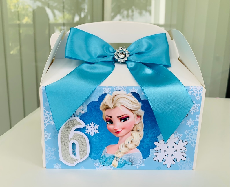 K&ouml;nnte beinhalten: Wei&szlig;e Geschenkbox mit hellblauer Schleife und Strassverzierung. Die Box zeigt Elsa aus Frozen, Schneeflocken und die Zahl 6. Ideal f&uuml;r eine Geburtstagsfeier.