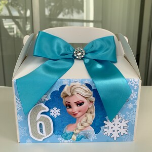 K&ouml;nnte beinhalten: Wei&szlig;e Geschenkbox mit hellblauer Satinschleife und Strassverzierung. Die Box zeigt ein Cartoon-Bild von Elsa aus Frozen, Schneeflocken und die Zahl 6 in silbernem Glitzer.