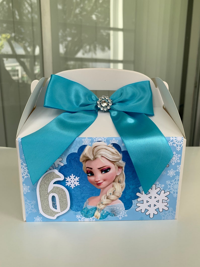 K&ouml;nnte beinhalten: Wei&szlig;e Geschenkbox mit hellblauer Satinschleife und Strassverzierung. Die Box zeigt ein Cartoon-Bild von Elsa aus Frozen, Schneeflocken und die Zahl 6 in silbernem Glitzer.