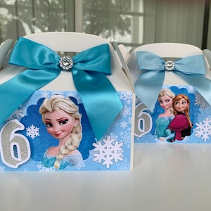 K&ouml;nnte beinhalten: Zwei wei&szlig;e Gastgeschenkboxen mit hellblauen Schleifen und Strassverzierungen. Jede Box zeigt ein anderes Bild von Figuren aus dem Film Frozen, mit Schneeflocken und der Zahl 6.