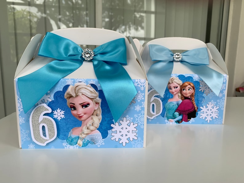 K&ouml;nnte beinhalten: Zwei wei&szlig;e Gastgeschenkboxen mit hellblauen Schleifen und Strassverzierungen. Jede Box zeigt ein anderes Bild von Figuren aus dem Film Frozen, mit Schneeflocken und der Zahl 6.
