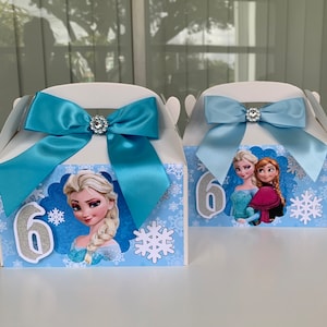 K&ouml;nnte beinhalten: Zwei wei&szlig;e Geschenkboxen mit hellblauen Schleifen und Strassverzierungen. Jede Box zeigt Charaktere aus dem Film "Die Eisk&ouml;nigin", Schneeflocken und die Zahl 6 in silbernem Glitzer.